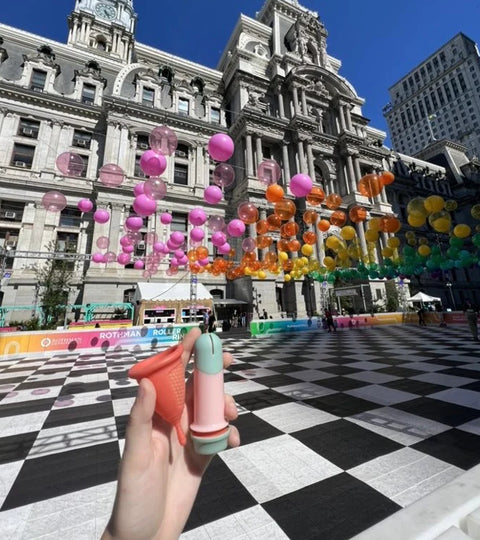 Sunny period cup and reusable applicator in front of building celebrating pride month with rainbow balloons